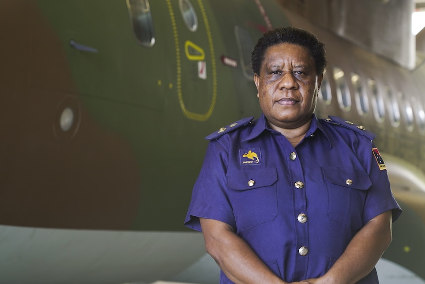 A Black woman stands next to a plane holding her arms in front of her.