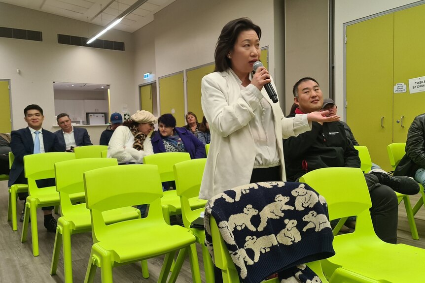 A woman in a small community hall audience speaks into a microphone. 