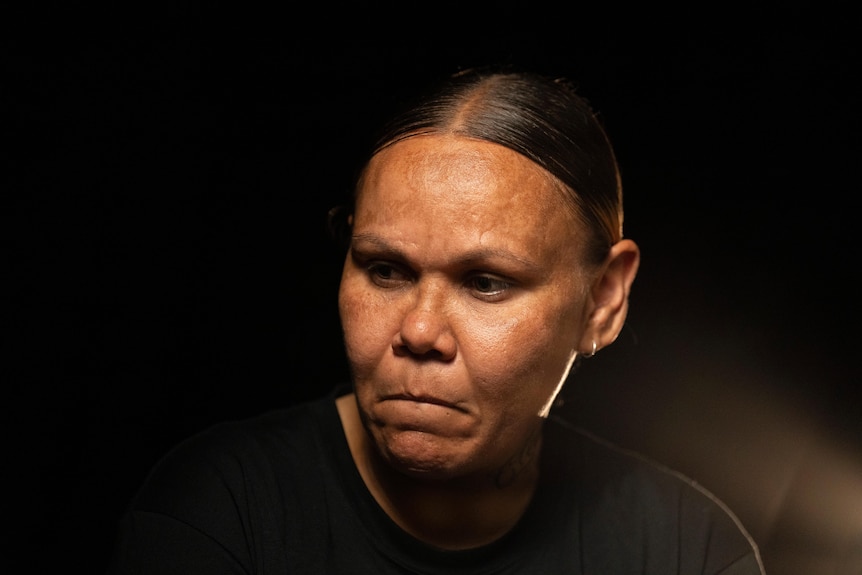 An Indigenous lady looks to the side with a solemn expression pictured against a black background