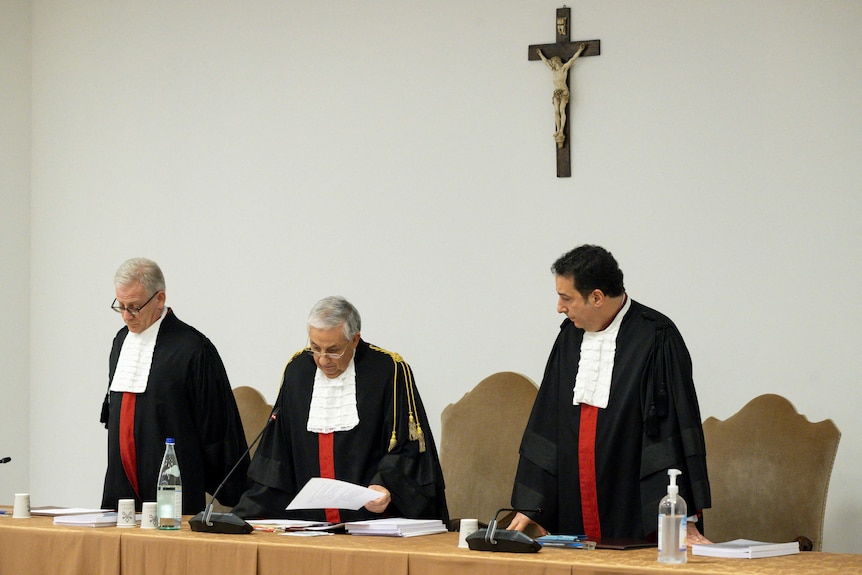 three judges stand while the one in the middle reads out a verdict. there is a jesus on a cross hanging behind them on the wall