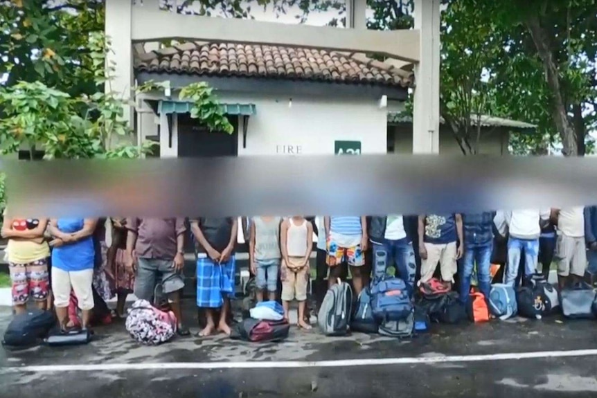 A group of people stand in a horizontal line in front of a building with their faces blurred.