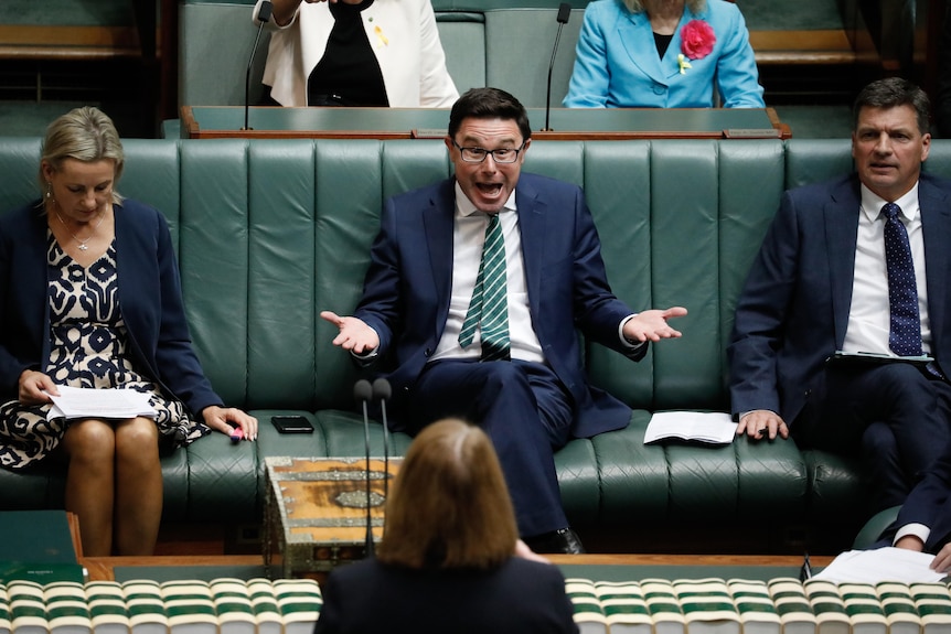 A dark-haired, bespectacled man sits in parliament reacting in a gregarious fashion to a speech.