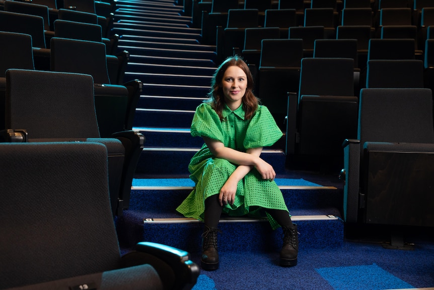 Laura Murphy, a brunette white woman in her 30s, wears a bright green dress and sits on the steps of a theatre seating bank.