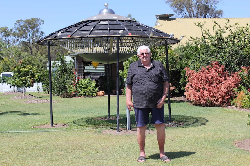 A man stands in front of a large UFO sculpture in his front yard