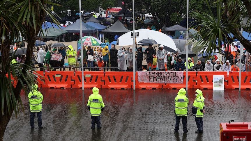 Four yellow vested police officers stand opposite a crowd of protesters on a rainy day at government building