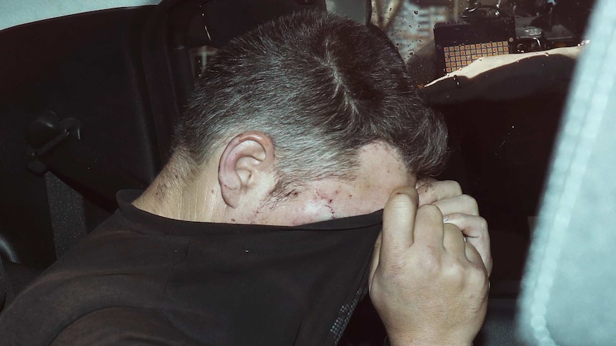 Jonathan Dick covers his face with a black t-shirt as he suits in the back passenger seat of a car.
