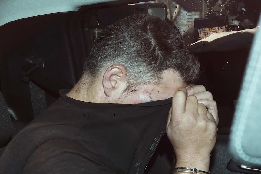 Jonathan Dick covers his face with a black t-shirt as he sits in the back passenger seat of a car.