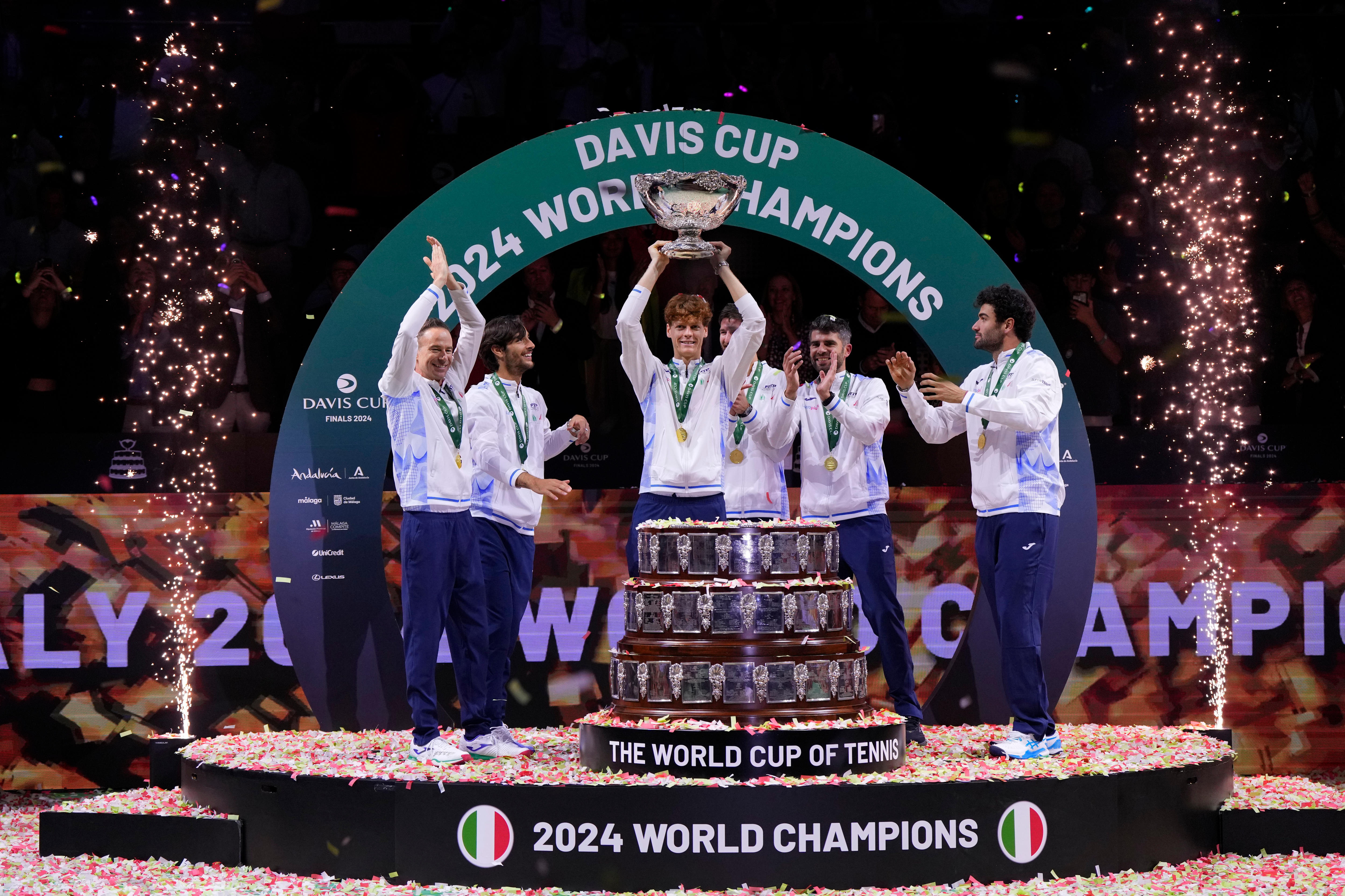 A group of Italian tennis players stand on a podium with the Davis Cup trophy as confetti is fired at either side of them.