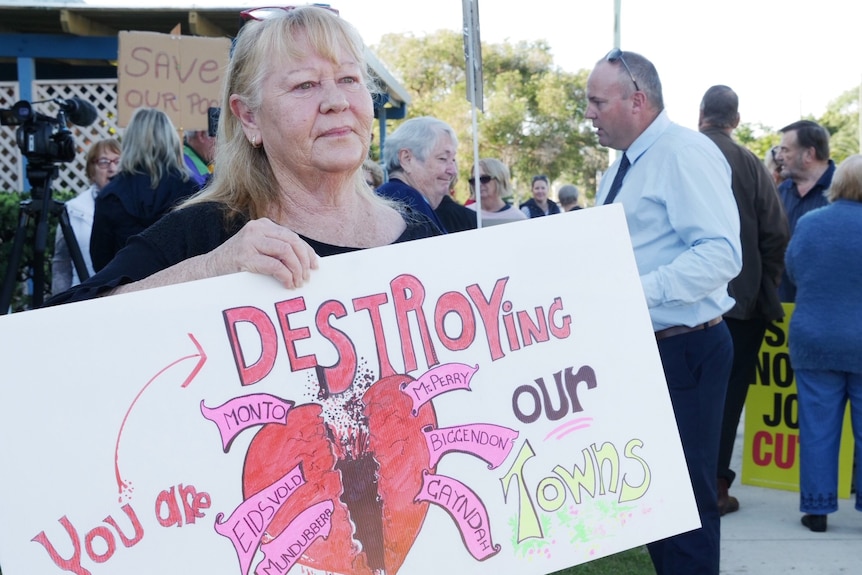 An older woman looks concerned as she holds a sign reading "you are destroying our town".