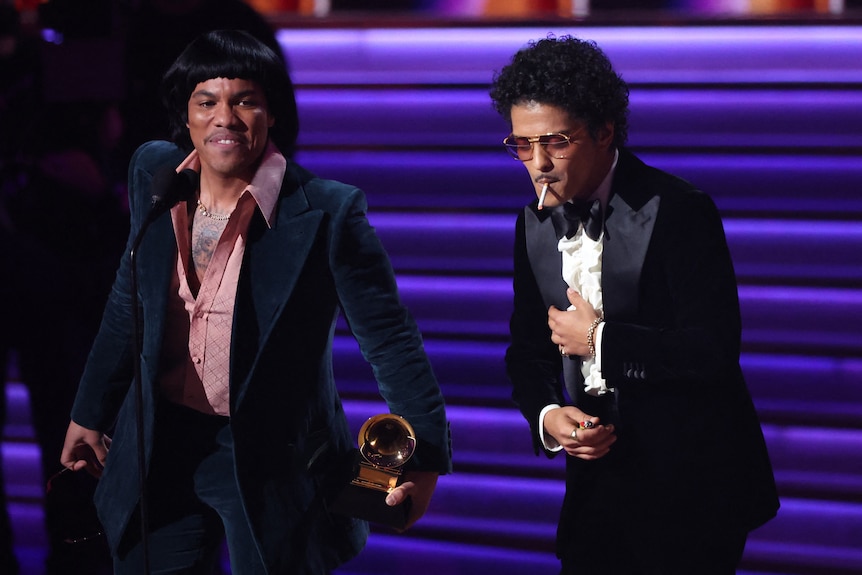 bruno mars takes a drag of a cigarette while anderson .paak accepts a grammy on stage 