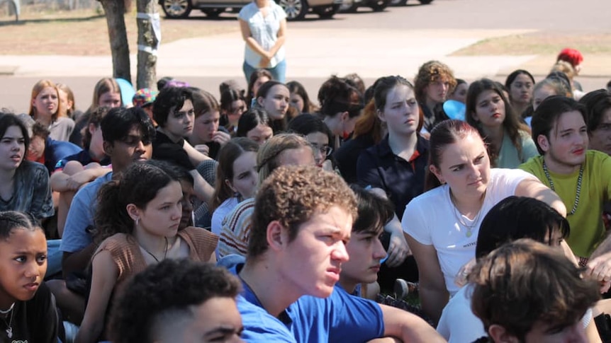 A crowd of high school students sitting on the ground listening to someone out of shot.