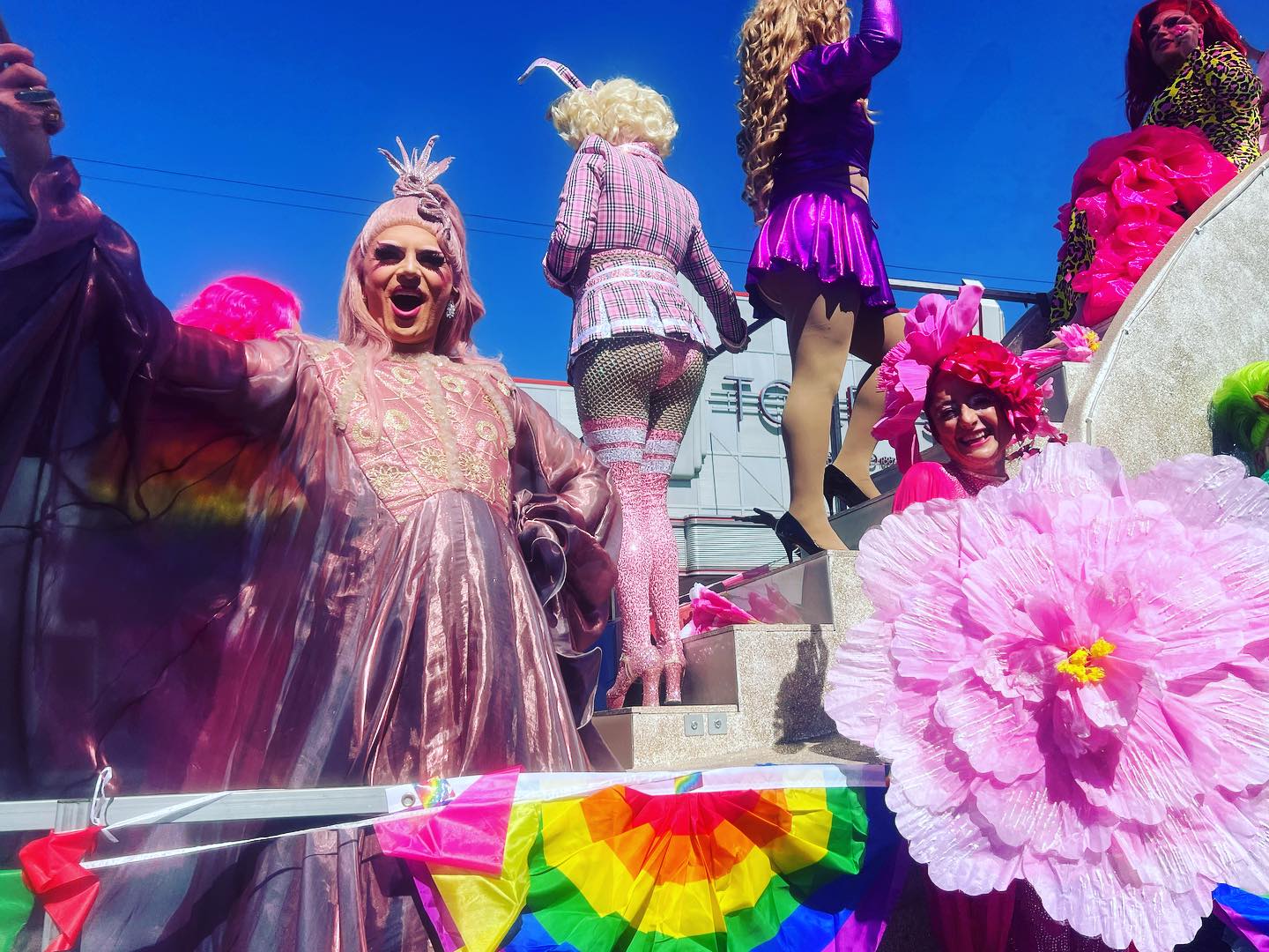 A group of drag queens dressed up in colourful outfits standing on and next to a parade float