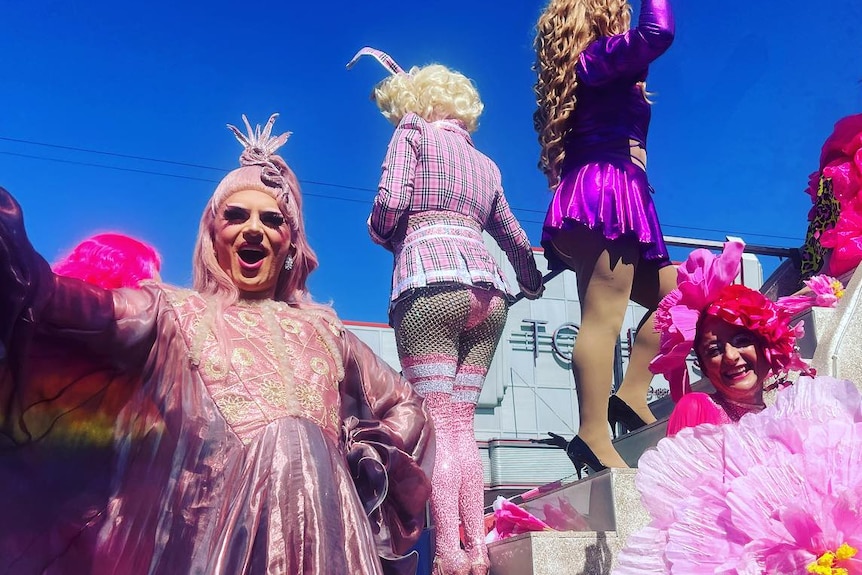 A group of drag queens dressed up in colourful outfits standing on and next to a parade float
