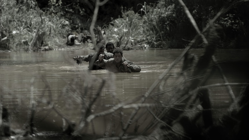 A desaturated image of three Black men dressed in tattered clothing wading across a mucky body of water. 
