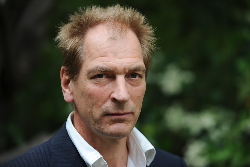 A closeup headshot of Julian Sands looking at the camera with messy hair and a black jacket and white collar
