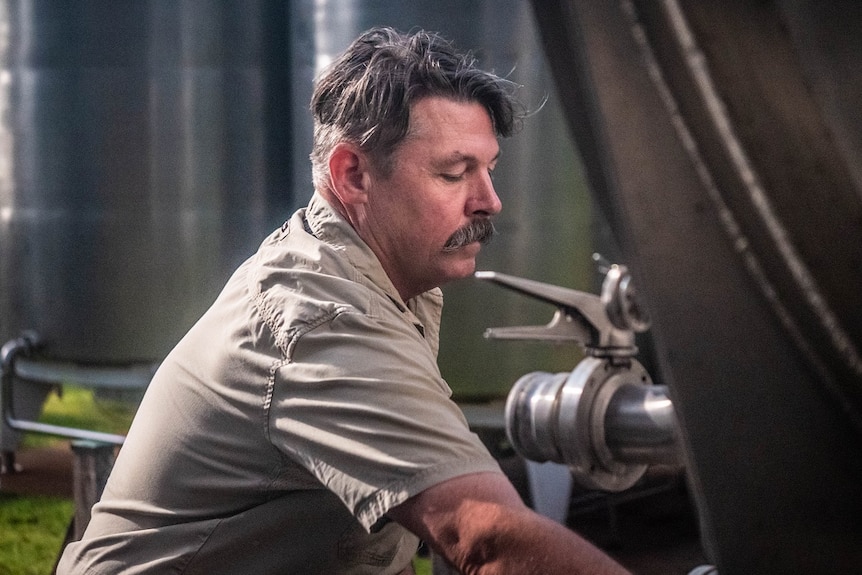 A man with a thick moustache kneels before a large tap, surrounded by stainless steel tanks.