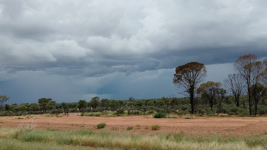 An outback landscape with red dirt and large dark storm clouds rolling overhead.