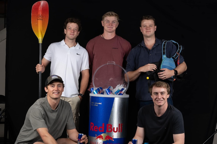 Five young men, three standing, two sitting with a about a metre-high can of Red Bull between. One holds a red paddle.