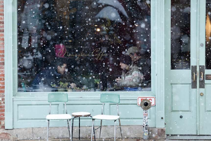 People can be seen through a window sitting in a cafe as snow falls outside
