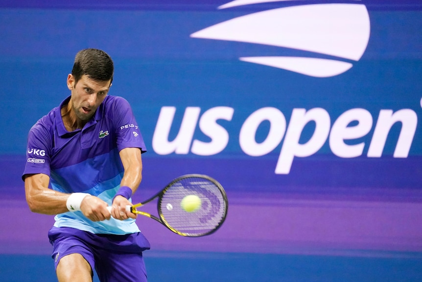 Novak Djokovic plays a two-handed backhanded in front of a blue wall with US Open written on it.
