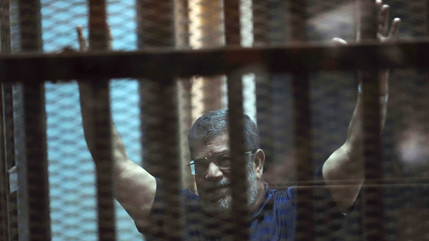 Former Egyptian President Mohammed Morsi waves to supporters in a blue polo shirt with both hands in the air.