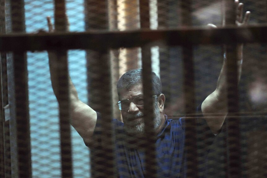 Former Egyptian President Mohammed Morsi waves to supporters in a blue polo shirt with both hands in the air.