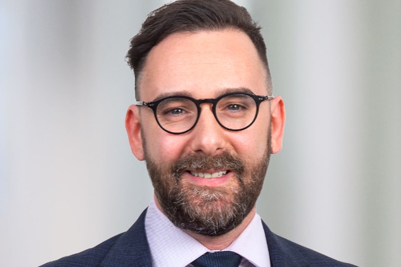 A professional headshot of Will Stidston wearing a suit and glasses.