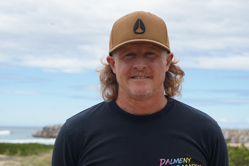A man with strawberry long hair and a hat smiles at a camera with waves in the background.