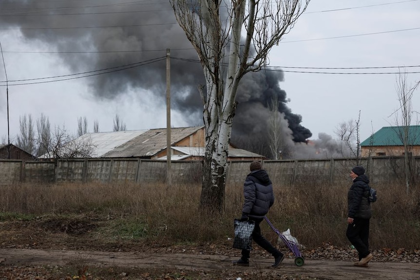 Two people walk past a fence, above it smoke billows out from a building that has been struck by a missile 