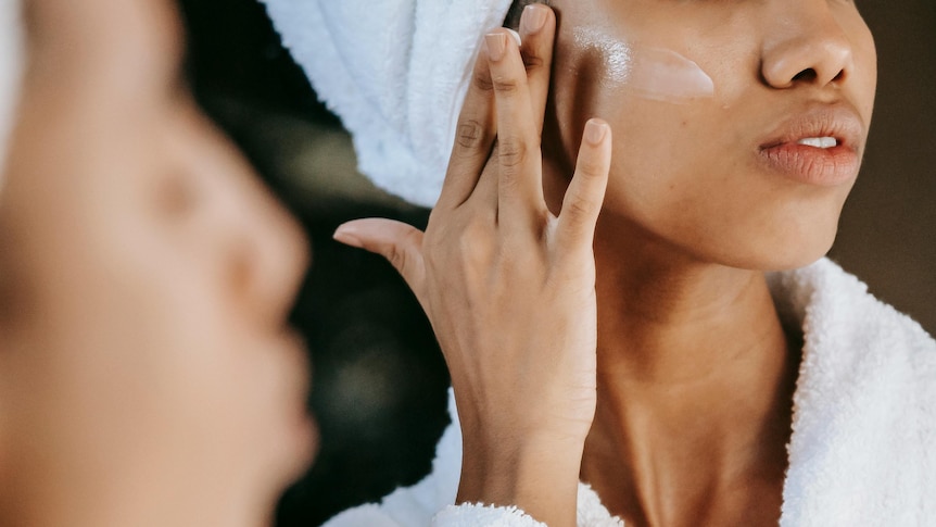Woman applies face cream while looking into a mirror. Her hair is wrapped in a towel and she is wearing a bath robe. 