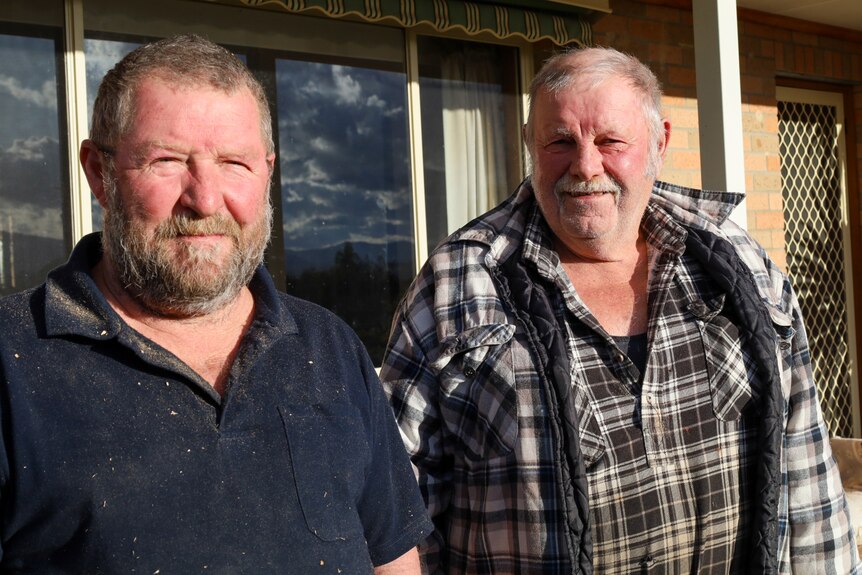Two older men smile into the sun on the front verandah of a house.