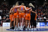 The Canberra Capitals players stand in a circle in the middle of a basketball court, in front of a crowd.