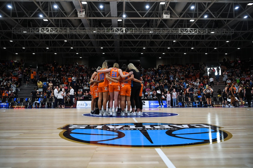 The Canberra Capitals players stand in a circle in the middle of a basketball court, in front of a crowd.