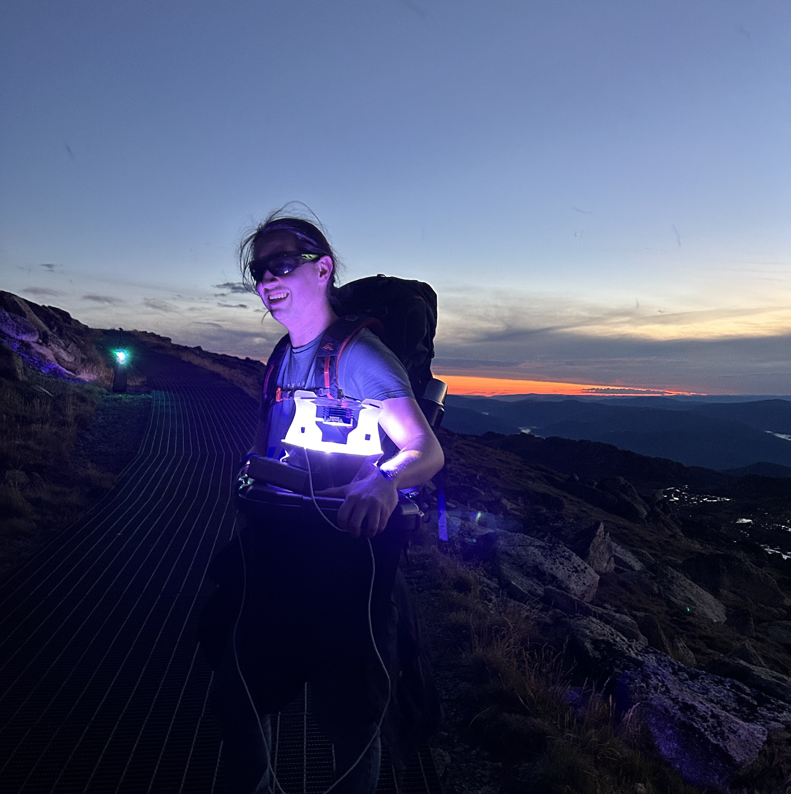 A man in a t-shirt carrying a large backpack and an illuminated lantern, standing on a mountain path