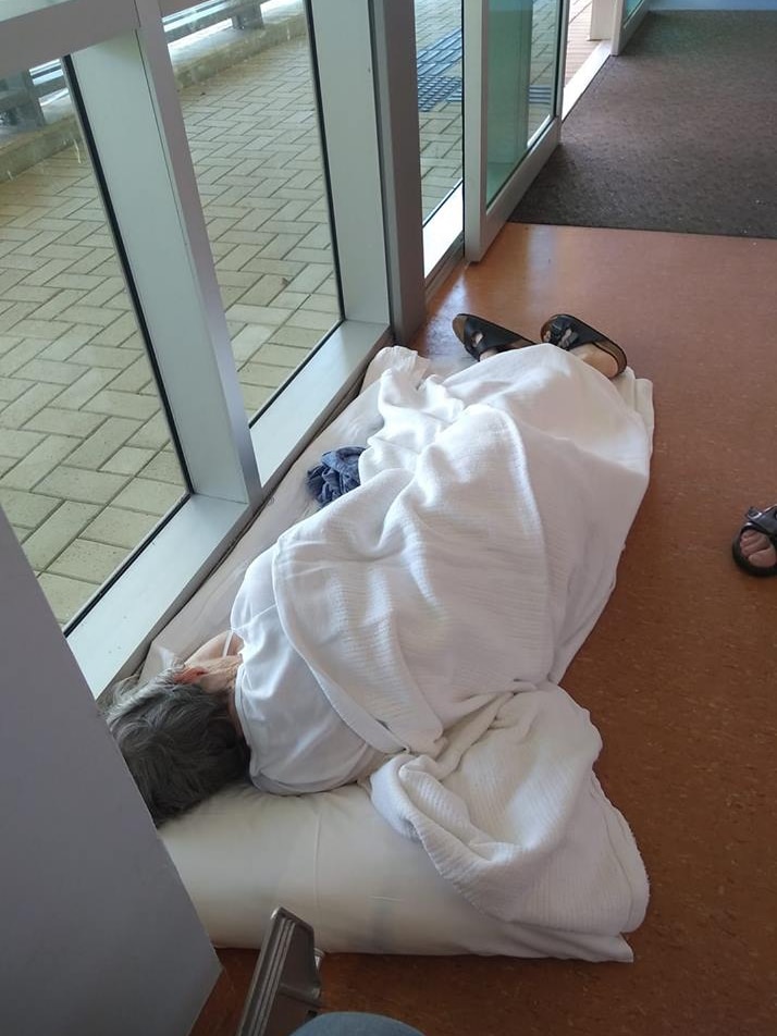 A woman lies on the floor of a hospital corridor covered in a white blanket.