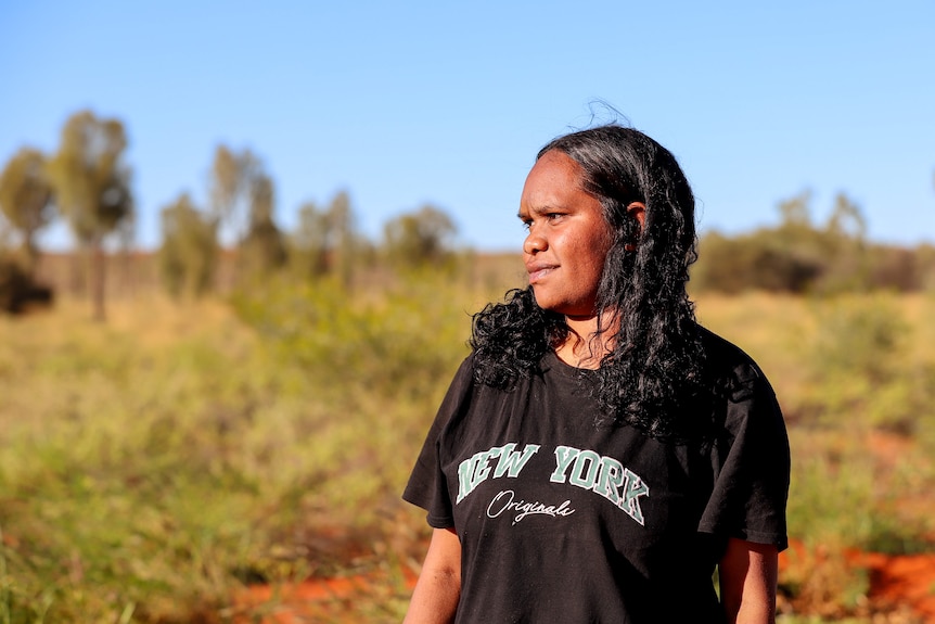 Young Aboriginal woman stands looking sideways on red dirt wtih blue sky and scrub behind her