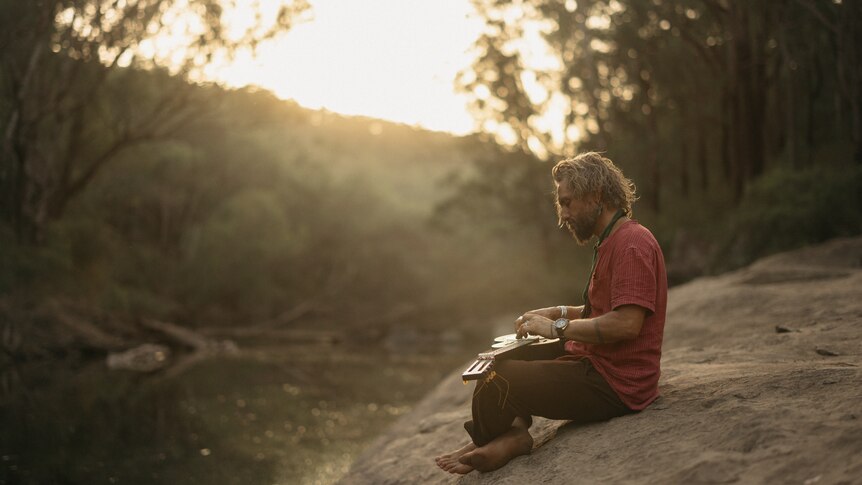 Australian musician John Butler sits on a rock with his guitar overlooking the Australian bush