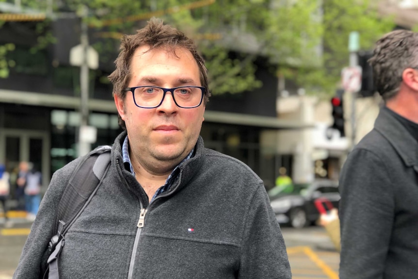 A man with glasses, brown hair and a grey top looks calmly at the camera.