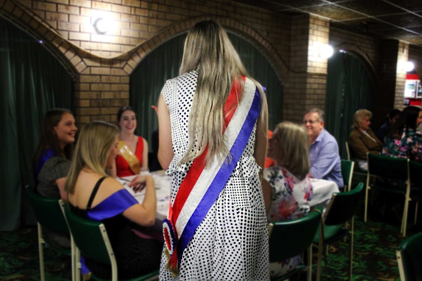 Back view of a woman wearing a winning sash approaching a table with other sash-wearing young women.