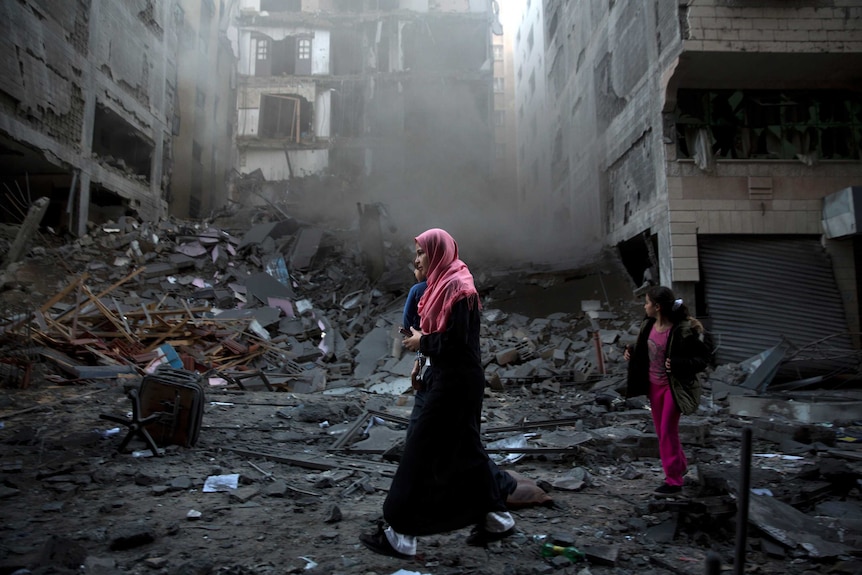 Palestinian people walk among the rubble of a destroyed building.