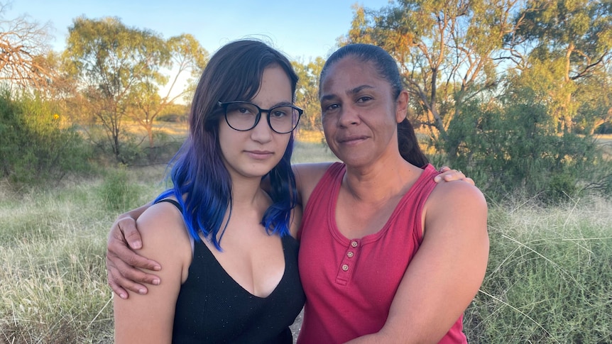 An Indigenous mother stands with her arms around her teenage daughter.