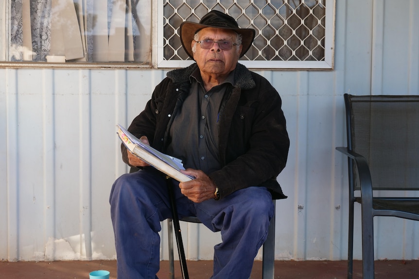 Man holding documents sits in front of a blue tin house.