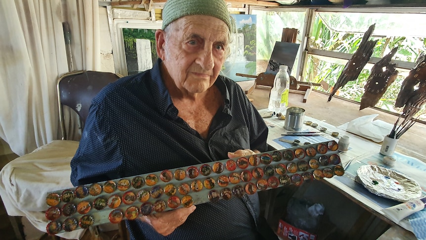 An unsmiling elderly man holds up a long metal bar covered in painted bottle caps, easel, brushes behind him.