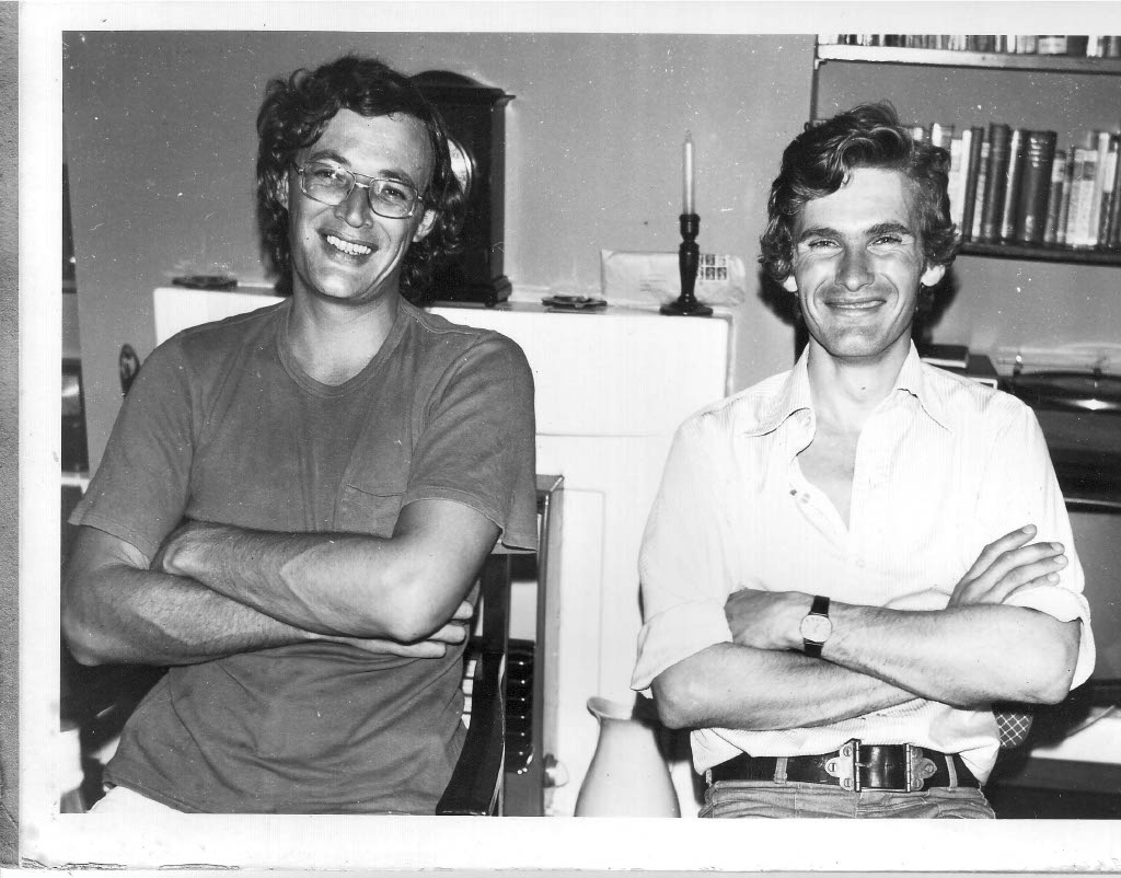 Man sits smiling in t-shirt and glasses. He has dark hair.