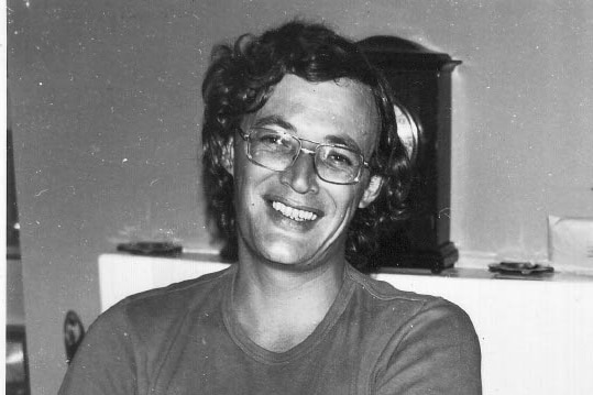 Man sits smiling in t-shirt and glasses. He has dark hair.