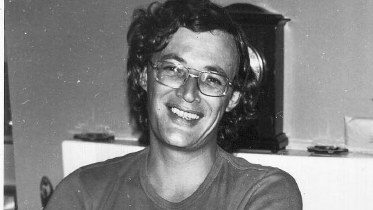 Man sits smiling in t-shirt and glasses. He has dark hair.