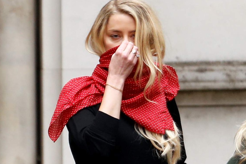 A woman with blond hair holds a red scarf across her face.