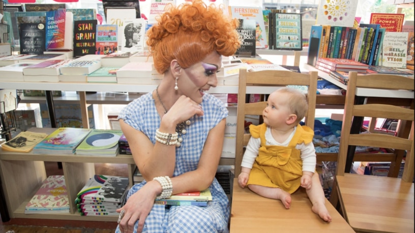 a drag performer with a bright wig is smiling at a toddler in a bookshop 