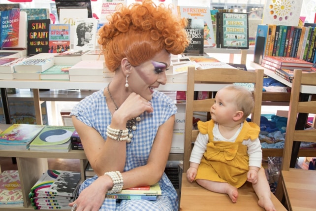 a drag performer with a bright wig is smiling at a toddler in a bookshop 