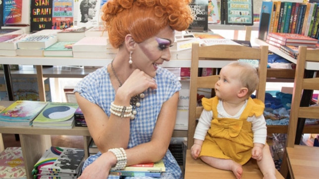 A drag performer sits on a chair beside a small child